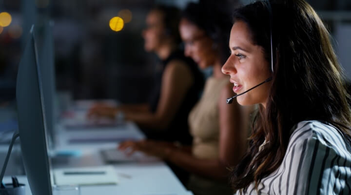 Customer support agents work late while wearing headsets at computer stations, showing how limited customer service hours are impacting your business.