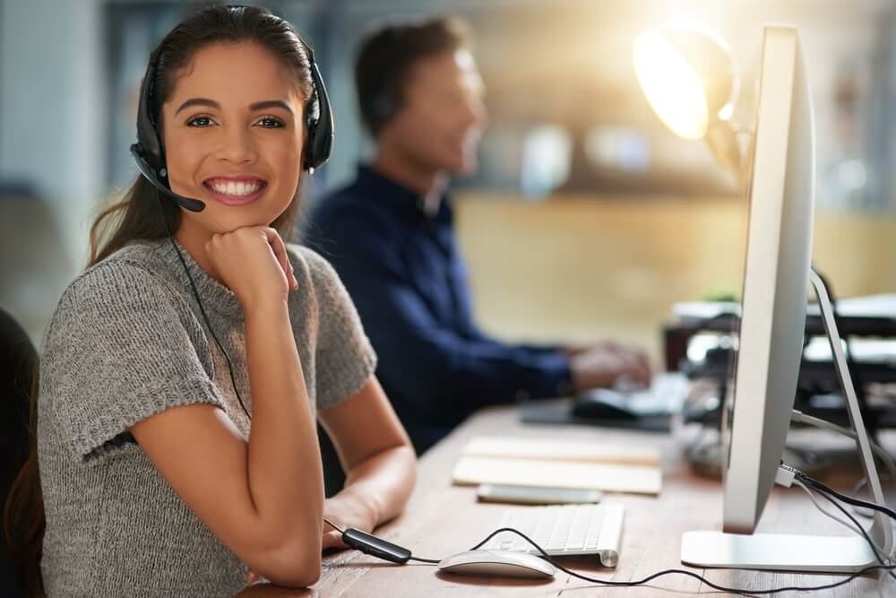 Customer service agent smiling at her desk while wearing a headset, showing how limited customer service hours are impacting your business.