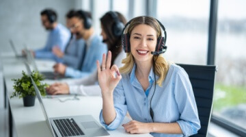smiling female customer support agent wearing a headset makes an ok hand gesture at her desk while coworkers work in the background, showing the benefits of outsourcing customer support.