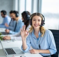 smiling female customer support agent wearing a headset makes an ok hand gesture at her desk while coworkers work in the background, showing the benefits of outsourcing customer support.
