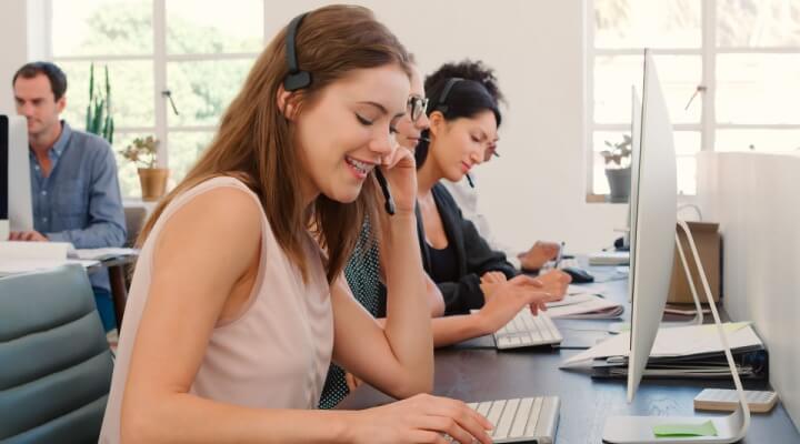 Multi-Cultural Female Business Team Wearing Headsets Working In Busy Customer Service Department.