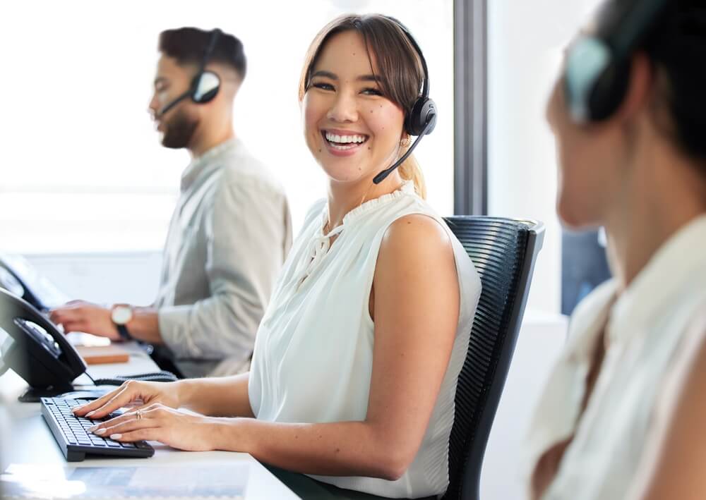 Smiling bilingual, bicultural call center worker looks at the camera while her colleagues work around her - best BPO partner.