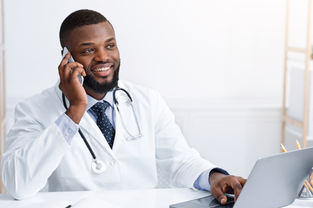 A male doctor talks on the phone while sitting at a desk in front of his laptop.