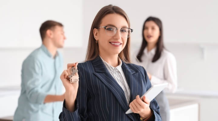 Woman real estate agent holds keys and paperwork, in the background is couple with boxes who just bought a new home.