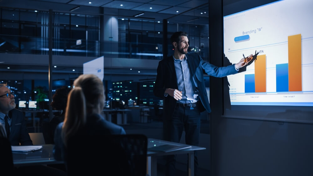 Conference business meeting presentation: chief executive officer shows data to diverse group of investors, businesspeople. projector screen shows graphs, product sales growth, e-Commerce analysis, how BPO helps drive vertical growth.