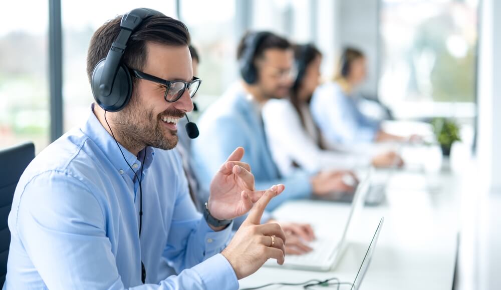 Professional customer support agent man counting on fingers while assisting customers at call center office, with colleagues in the background, nearshoring and onshoring BPO.