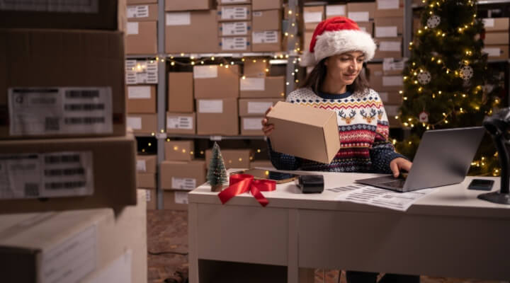 Employee in retail shop's warehouse in Santa hat needs holiday customer support outsourcing. Small Business Owner working on laptop, preparing and packing parcels for delivery in holidays.