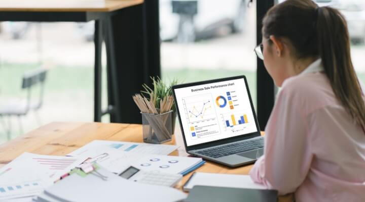 Woman entrepreneur with glasses reviews growth charts and spreadsheets on her laptop, considering whether to outsource small business bookkeeping.