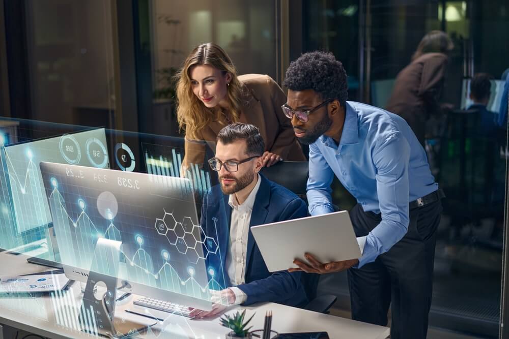 Three coworkers analyze data on a desktop monitor with futuristic AI-style overlays while one holds a laptop in a late-night office, highlighting BPO labor trends and tech-driven decision-making.