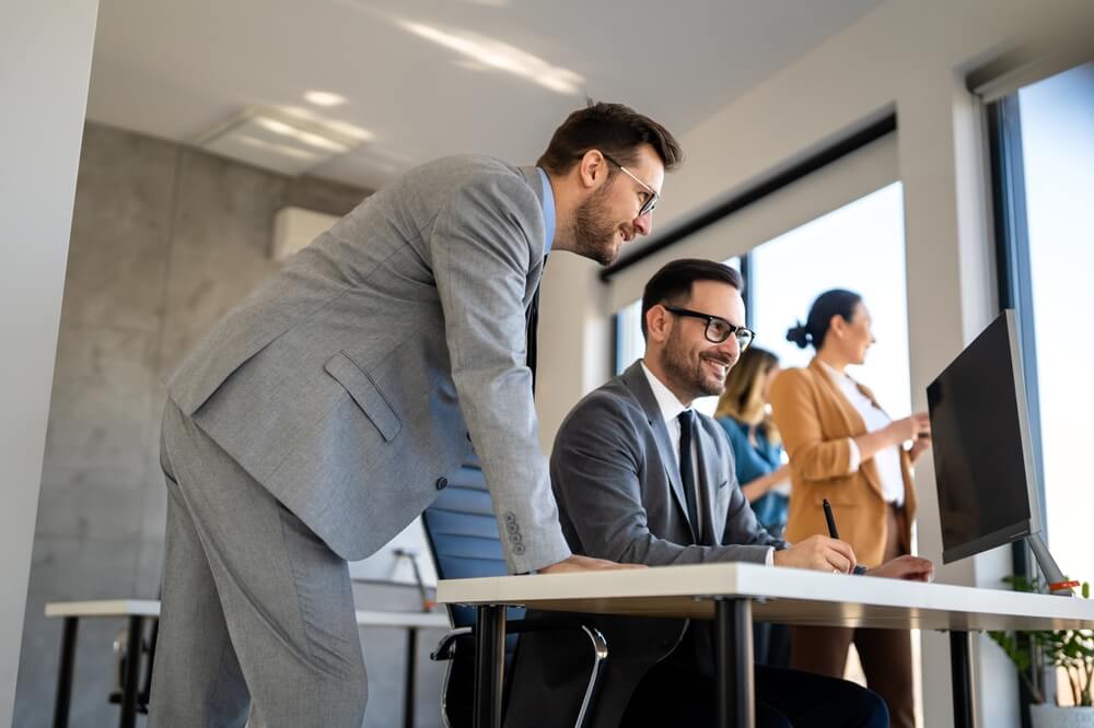 Two business professionals in suits collaborate at a desktop computer in a modern office while teammates work in the background, illustrating hyperautomation in BPO.