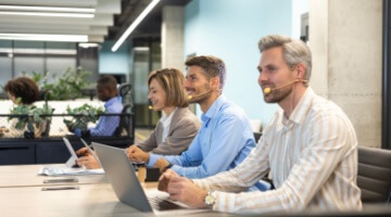 Outsourced call center agents wearing headsets working at laptops in a modern open office, smiling and assisting customers while seated in a row.