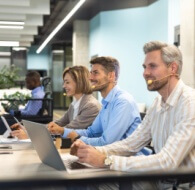 Outsourced call center agents wearing headsets working at laptops in a modern open office, smiling and assisting customers while seated in a row.