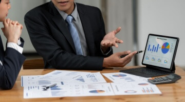 Business meeting discussing BPO contract with professional presenting charts and graphs on a tablet, with reports on the table.