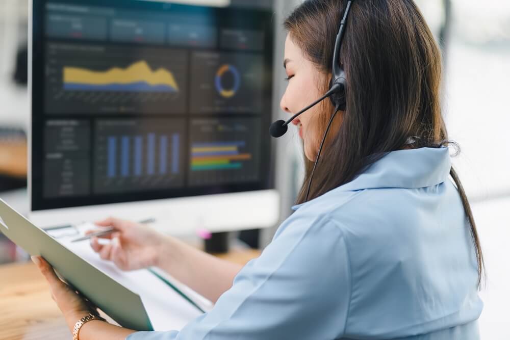 Customer support agent wearing a headset reviews a report on a clipboard while monitoring performance dashboards on a computer for Back-Office BPO Services.