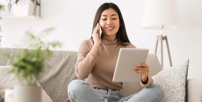 Young woman sitting on a couch while making a call and using a tablet.